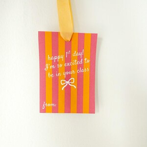 Pu&ograve; includere: Un'etichetta regalo rettangolare con strisce verticali arancioni e rosa. L'etichetta riporta il testo "happy 1st day! I'm so excited to be in your class" e un fiocco bianco. Un nastro giallo &egrave; attaccato in alto.