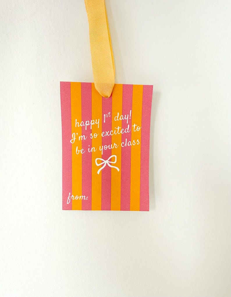 Pu&ograve; includere: Un'etichetta regalo rettangolare con strisce verticali arancioni e rosa. L'etichetta riporta il testo "happy 1st day! I'm so excited to be in your class" e un fiocco bianco. Un nastro giallo &egrave; attaccato in alto.