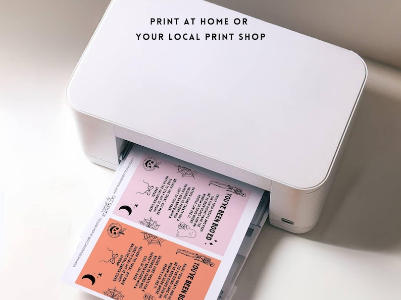 May include: A white printer is shown printing pink and orange Halloween-themed cards. The cards have black illustrations of ghosts, spiders, and pumpkins. Text on the cards reads "YOU'VE BEEN BOOED" and "PRINT AT HOME OR YOUR LOCAL PRINT SHOP."