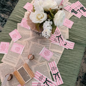 Pode incluir: Uma decora&ccedil;&atilde;o de mesa com um arranjo floral de rosas brancas e pequenas flores brancas. A pe&ccedil;a central fica em uma base de p&aacute;ginas de livros abertas. Marcadores listrados rosa e branco e cart&otilde;es rosa com texto est&atilde;o espalhados.