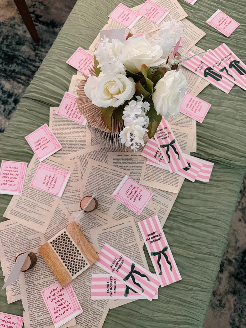 Pode incluir: Uma decora&ccedil;&atilde;o de mesa com um arranjo floral de rosas brancas e pequenas flores brancas. A pe&ccedil;a central fica em uma base de p&aacute;ginas de livros abertas. Marcadores listrados rosa e branco e cart&otilde;es rosa com texto est&atilde;o espalhados.