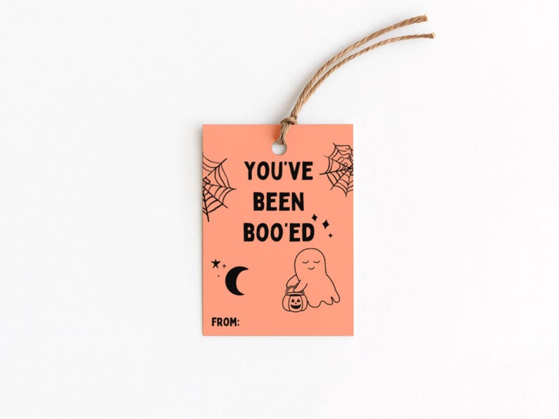 May include: A rectangular orange gift tag with the text "YOU'VE BEEN BOO'ED" in black. The tag features a ghost holding a pumpkin, spiderwebs, a crescent moon, and stars. A brown string is attached at the top.