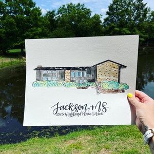Puede incluir: Pintura a la acuarela de una casa con el texto "Jackson, MS 2615 Highland Place Drive". La casa tiene un tejado negro, ladrillo y revestimiento gris, y ventanas te&ntilde;idas de azul. La pintura se sostiene frente a un estanque y &aacute;rboles.
