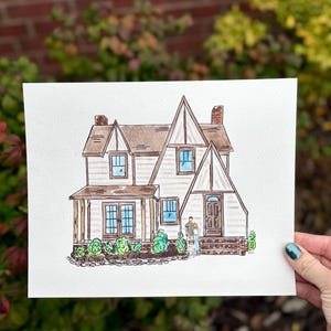 Puede incluir: Pintura a la acuarela de una casa con tejado marr&oacute;n, revestimiento blanco y cristales azules. La obra representa a una pareja de pie frente a la puerta. Arbustos verdes y un fondo de ladrillo completan la escena.