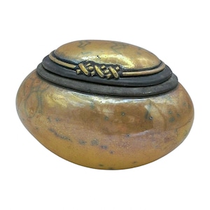 Puede incluir: Caja decorativa ovalada con tapa, acabado metálico bronce. Una banda oscura rodea el centro. La tapa está adornada con detalles dorados y un cierre decorativo dorado.