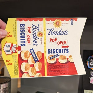 May include: A vintage box of Borden's Pop Open Biscuits. The box is red, white, and blue with a picture of biscuits on the front. The text on the box reads "Borden's Pop Open Biscuits Enriched".