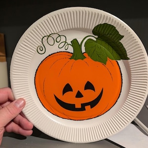 May include: White paper plate with a smiling orange pumpkin with a green stem and leaf. The pumpkin has a black face with a wide smile and two eyes.