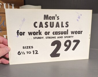 Op de afbeelding: Een wit bord met zwarte tekst dat heren casuals voor werk of vrijetijdskleding adverteert. Het bord zegt "Men's Casuals for work or casual wear... Sturdy, Strong and Sporty" en "Sizes 6 1/2 to 12" en "297".