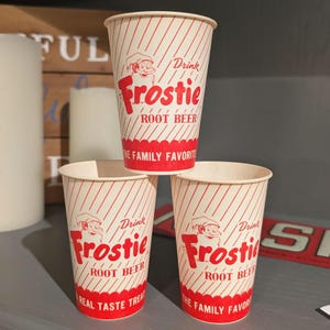 May include: Three red and white striped paper cups with the words "Drink Frostie Root Beer" printed on them. The cups are stacked on top of each other.