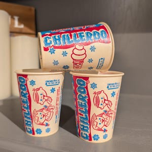 Puede incluir: Tres vasos de helado Chilleroo vintage. Los vasos son de color beige con gráficos rojos y azules, incluyendo la palabra "Chilleroo", copos de nieve y un cono de helado. Un vaso está tumbado de lado y dos están de pie.