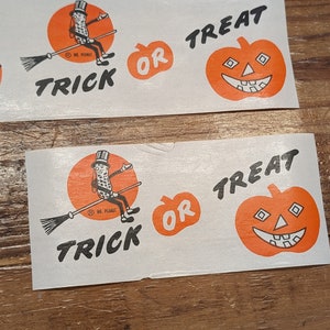 K&ouml;nnte beinhalten: Zwei wei&szlig;e rechteckige Aufkleber mit Halloween-Motiven. Einer zeigt Mr. Peanut auf einem Besen mit dem Wort "TRICK" darunter. Der andere hat einen orangefarbenen K&uuml;rbis mit "OR TREAT".