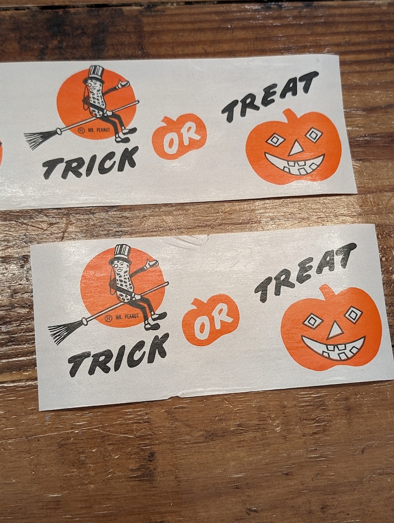 K&ouml;nnte beinhalten: Zwei wei&szlig;e rechteckige Aufkleber mit Halloween-Motiven. Einer zeigt Mr. Peanut auf einem Besen mit dem Wort "TRICK" darunter. Der andere hat einen orangefarbenen K&uuml;rbis mit "OR TREAT".