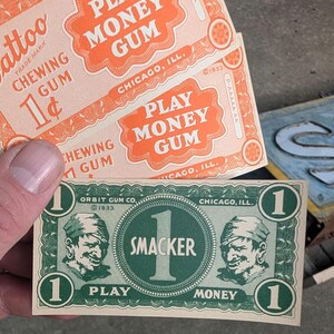 K&ouml;nnte beinhalten: Nahaufnahme von Vintage "Play Money Gum" und "Smacker" Spielgeld. Die orangefarbenen und wei&szlig;en Kaugummipapierchen tragen die Aufschrift "Chewing Gum" und "1&cent;". Das gr&uuml;ne und wei&szlig;e Spielgeld zeigt einen Piraten und den Text "Play Money".