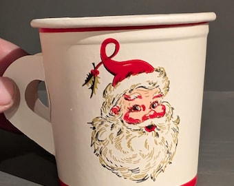 Puede incluir: Vaso de papel blanco con un borde rojo y un asa. El vaso presenta una ilustración vintage de Papá Noel con un gorro rojo y una barba blanca.