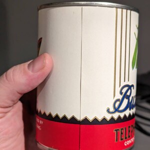 May include: A white metal can with a red, black, and gold label. The label has the text "TELEPHONE" and a stylized blue cursive word "Butter" with a green butterfly graphic.