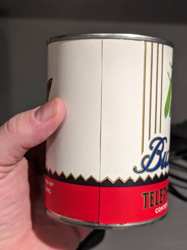 May include: A white metal can with a red, black, and gold label. The label has the text "TELEPHONE" and a stylized blue cursive word "Butter" with a green butterfly graphic.