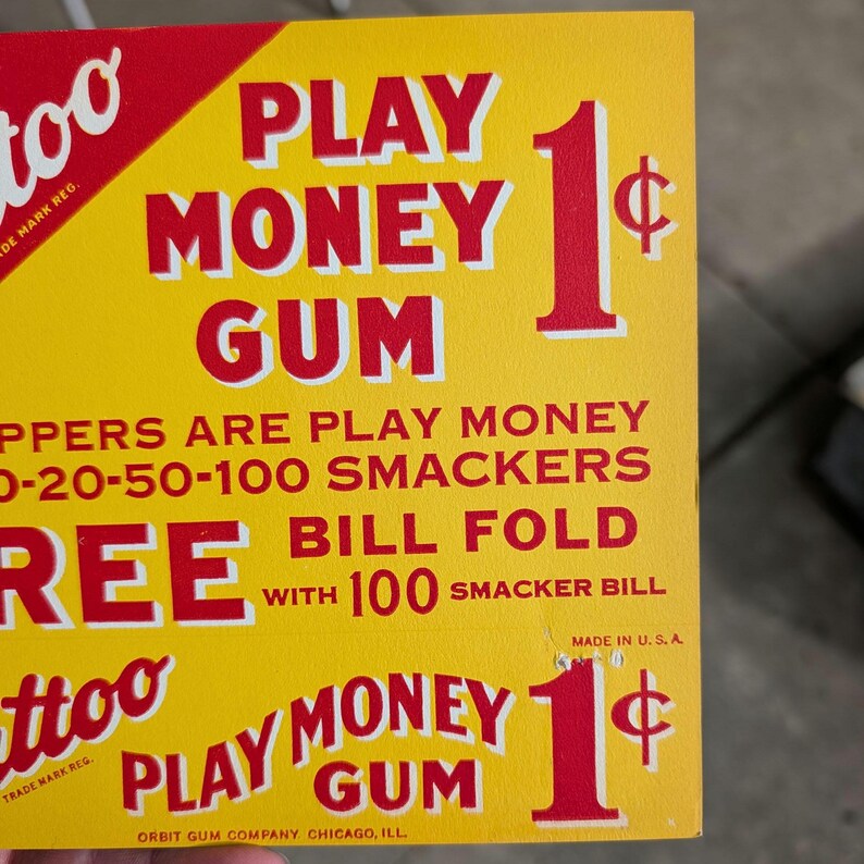 K&ouml;nnte beinhalten: Ein gelb-rotes Vintage-Schild f&uuml;r "Play Money Gum" mit einem gro&szlig;en roten "1&cent;"-Symbol. Das Schild enth&auml;lt Text &uuml;ber den Kaugummi und den Satz "Made in U.S.A."