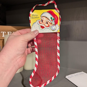 Puede incluir: Una media roja y blanca a rayas de tejido de malla con una tarjeta de Papá Noel unida a la parte superior. La tarjeta dice "Feliz Navidad" y presenta un Papá Noel de dibujos animados con barba blanca y gorro rojo.