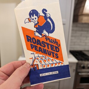 Peut inclure: Un sac en papier blanc avec un design bleu et orange repr&eacute;sentant un singe de dessin anim&eacute; tenant une queue. Le sac indique "Fresh Roasted Peanuts" et "Net Wt. 1 oz. or over".