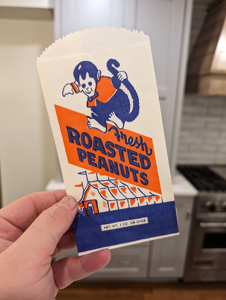 Peut inclure: Un sac en papier blanc avec un design bleu et orange repr&eacute;sentant un singe de dessin anim&eacute; tenant une queue. Le sac indique "Fresh Roasted Peanuts" et "Net Wt. 1 oz. or over".