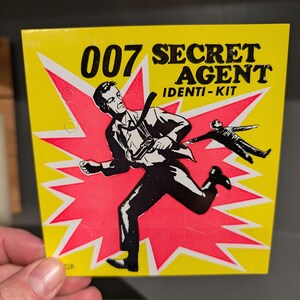 May include: A yellow and red card with the text "007 Secret Agent Identi-Kit" and a black and white illustration of a man running with a gun.