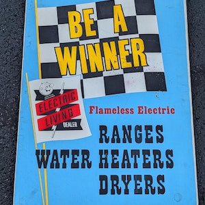 May include: A blue and white sign with a checkered flag and the text "Be a Winner". Below the flag is a red and white logo that says "Electric Living Dealer". The sign also says "Flameless Electric Ranges Water Heaters Dryers".