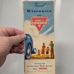 May include: Vintage travel brochure for Wisconsin with the Conoco logo. The brochure features a family illustration, a classic car, and handwritten notes. The text includes "Travel Wisconsin with CONOCO" and "TOURAIDE".