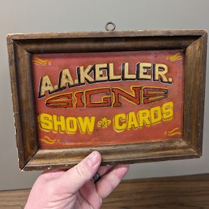 May include: A vintage wooden framed sign with a red background and gold lettering. The sign reads "A.A. Keller Signs Show Cards".