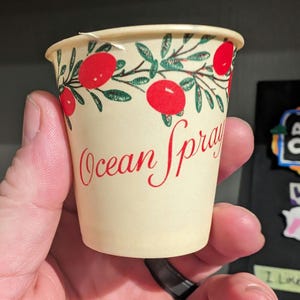 Puede incluir: Un vaso de papel blanco con un diseño rojo y verde que presenta una vid frondosa con bayas rojas y el texto "Ocean Spray" en letras rojas.