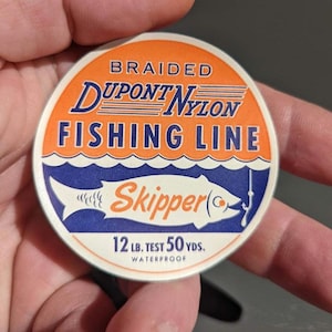May include: Round spool of vintage fishing line with orange, blue, and white colors. The text reads "Braided Dupont Nylon Fishing Line" and "Skipper". It also indicates 12 lb test and 50 yds.