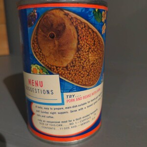 May include: A blue and orange can of pork and beans with ham. The can has a picture of the beans and ham on the front. The text on the can reads "MENU SUGGESTIONS TRY... PORK AND BEANS WITH HAM A tasty, easy to prepare, main dish suitable for family and Sunday night suppers. Serve with a tossed green salad and coffee. Truly an inexpensive meal for a thrift minded housewife. SIZE OF THIS CAN... NO. 1 - CUPFULS CONTENTS... 11 OZS. AVD. - SERVES 2-3"