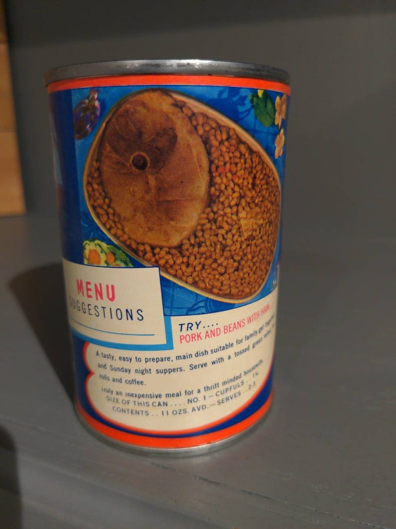May include: A blue and orange can of pork and beans with ham. The can has a picture of the beans and ham on the front. The text on the can reads "MENU SUGGESTIONS TRY... PORK AND BEANS WITH HAM A tasty, easy to prepare, main dish suitable for family and Sunday night suppers. Serve with a tossed green salad and coffee. Truly an inexpensive meal for a thrift minded housewife. SIZE OF THIS CAN... NO. 1 - CUPFULS CONTENTS... 11 OZS. AVD. - SERVES 2-3"