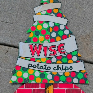 May include: A flat, tree-shaped sign featuring a pattern of red, orange, green, and white polka dots. Two white banners wrap around the tree, displaying the red text WISE and the black text potato chips. The base is a red brick pattern.