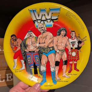 May include: A yellow paper plate featuring the WWF logo and illustrations of wrestlers Hulk Hogan, Roddy Piper, and others. The plate has a red center and is decorated with various wrestler names.