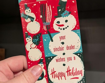Könnte beinhalten: Eine Vintage-Weihnachtsbox in Rot und Weiß mit Schneemann-Illustrationen. Auf der Box steht der Text "your sinclair dealer wishes you a Happy Holiday" in einer festlichen Schriftart. Die Box wird von einer Hand gehalten.