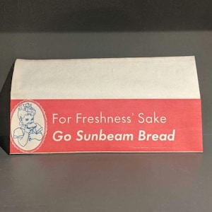 Op de afbeelding: Vintage Sunbeam Brood verpakking. De verpakking heeft een rode band met witte tekst: "For Freshness' Sake Go Sunbeam Bread." Een blauw-witte illustratie van een etende baby staat aan de linkerkant.