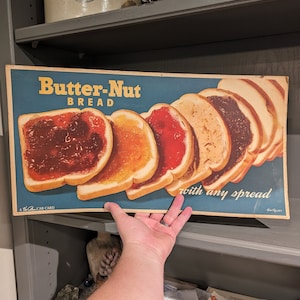 May include: Vintage advertisement for Butter-Nut Bread featuring a stack of sliced bread with different spreads. The text reads "Butter-Nut Bread" and "with any spread".