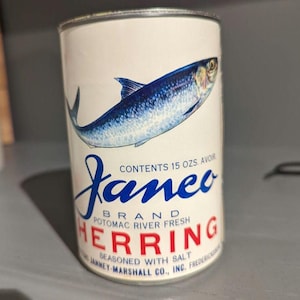 May include: A vintage can of Janeo Brand Potomac River Fresh Herring, seasoned with salt. The can is white with blue and red lettering and features a realistic illustration of a herring fish.