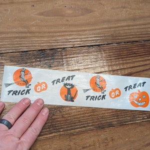 K&ouml;nnte beinhalten: Ein Vintage-Halloween-Papierstreifen mit orangefarbenen und schwarzen Illustrationen. Der Streifen zeigt eine schwarze Katze, einen K&uuml;rbis und eine Figur auf einem Besen mit den Worten "Trick or Treat".