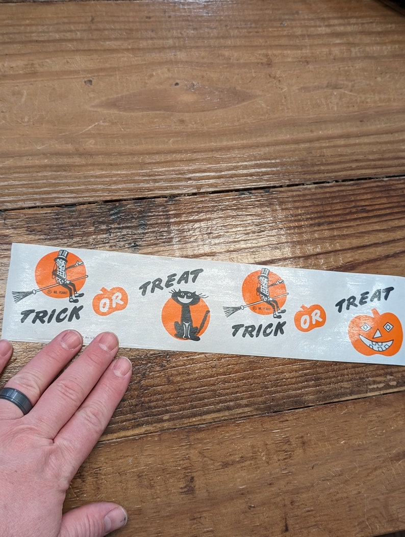 K&ouml;nnte beinhalten: Ein Vintage-Halloween-Papierstreifen mit orangefarbenen und schwarzen Illustrationen. Der Streifen zeigt eine schwarze Katze, einen K&uuml;rbis und eine Figur auf einem Besen mit den Worten "Trick or Treat".