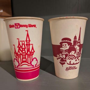 Puede incluir: Dos vasos de papel vintage. Uno presenta un diseño de castillo rosa con las palabras "Walt Disney World". El otro vaso tiene una banda granate con ilustraciones y las palabras "World Showcase".
