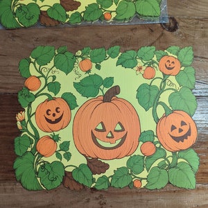 Op de afbeelding: Een groene en gele Halloween-placemat met een wijnstokpatroon en oranje pompoenen met jack-o'-lantern gezichten.