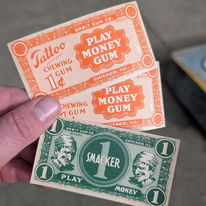 K&ouml;nnte beinhalten: Nahaufnahme von Vintage-Geldscheinen "Play Money Gum" und "Smacker Play Money". Die orangefarbenen Scheine tragen die Aufschrift "Tattoo Chewing Gum" und "Play Money Gum". Der gr&uuml;ne Schein tr&auml;gt die Aufschrift "Smacker Play Money". Alle Scheine stammen aus Chicago, Illinois.