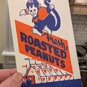 Peut inclure: Un sac en papier vintage avec un design bleu et rouge. Il repr&eacute;sente un singe de dessin anim&eacute; tenant une tente rouge et blanche ray&eacute;e avec le texte "Fresh Roasted Peanuts" et "Net Wt. 1 oz. or over".