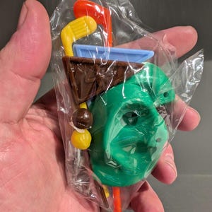 May include: A green plastic face mask with a brown, blue and yellow plastic accessory set. The set includes a golf club, a golf flag and a golf ball.