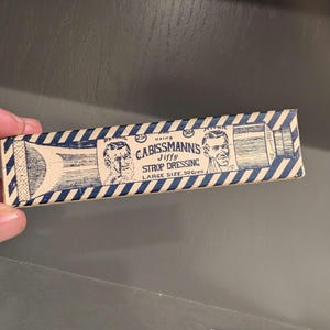 Può includere: Una scatola di cartone vintage per Cabissmann's Jiffy Strop Dressing. La scatola è marrone con strisce blu e bianche e presenta un'illustrazione in bianco e nero di un viso maschile. Il testo sulla scatola recita "Using Cabissmann's Jiffy Strop Dressing. Large Size. 50 cents."