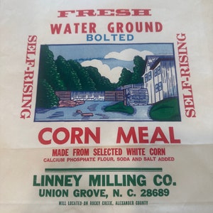 May include: A white paper bag with a green and blue illustration of a watermill. The bag is labeled "Fresh Water Ground Bolted Corn Meal" and "Linney Milling Co. Union Grove, N. C. 28689". The bag contains 10 pounds of cornmeal.