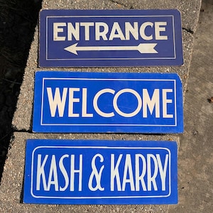 Könnte beinhalten: Drei rechteckige blaue Schilder mit weißem Text. Das obere Schild lautet "ENTRANCE" mit einem Pfeil nach links. Das mittlere Schild lautet "WELCOME". Das untere Schild lautet "KASH & KARRY".