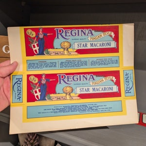 以下が含まれることがあります： 2つのヴィンテージRegina Star Macaroniラベル。ラベルは、"Regina Italian Style Star Macaroni"のテキストが入った赤と黄色のデザインです。青いドレスを着た女性がラベルに描かれています。