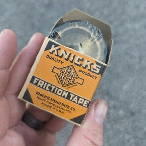 May include: A brown cardboard box with a roll of black friction tape inside. The box is labeled "KNICKS QUALITY PRODUCT" and "MEND-RITE FRICTION TAPE". The box also includes the company name and location: "KNICKS MEND-RITE CO. North Kansas City, Mo. MADE IN USA."